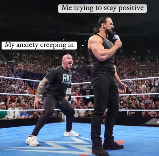 A photo from a WWE wrestling event shows two large men in the ring. The man on the right, Drew McIntyre, is standing upright and holding a microphone, looking serious. The text "Me trying to stay positive" is over his head. The man on the left, Randy Orton, is crouched low and screaming with his mouth wide open. The text "My anxiety creeping in" is over his head. The background is a packed crowd of fans watching the match.