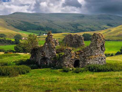 A high-angle landscape shot of the roofless, stone ruins of Lammerside Castle in Mallerstang, nestled in a lush green valley. The crumbling stone walls feature small arched doorways and jagged, uneven tops overgrown with grass and small bushes. Bright sunlight illuminates the verdant fields surrounding the structure, whilst sweeping fell sides rise dramatically in the background under a moody, cloud-filled sky casting soft shadows across the hills.
