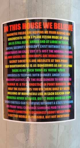 A sticker with rainbow-colored text, reading: IN THIS HOUSE WE BELIEVE MAGNETIC FIELDS ARE KEEPING ME FROM HAVING SEX JFK IS STILL ALIVE, ARMED AND AT LARGE IN TEXAS SOCIAL SECURITY WOULDN'T EXIST WITHOUT THE USSR CHEMTRAILS DO EXIST BUT IT'S JUST THE NORMAL POISON ALL OUR SOCIAL RELATIONSHIPS ARE MEDIATED BY IMAGES SECRET SOCIETIES ARE OBSELETE AT THIS POINT SELF RIGHTEOUSNESS IS AS DANGEROUS AS ANY WEAPON THERE IS NO SUCH THING AS 'OUTER' SPACE AMERICA IS TEEMING WITH HUNGRY, ANGRY GHOSTS MACROPLASTICS ARE THE REAL DANGER TO OUR HEALTH 'ALASKA' IS A TV SOUNDSTAGE IN POINT ROBERTS, WA HALF THE ELEVATORS YOU USE WERE BUILT BY NAZIS MILLIONS OF HOMONCULI LIVE IN EACH SILICON CHIP MKULTRA NEVER WORKED, BUT IT MADE GOOD AD COPY DRUG CARTELS CAN'T SURVIVE WITHOUT A STATE SPONSOR NO PROOF WILL CONVINCE SOMEONE PAID TO DISAGREE THERE'S SOMETHING DISHONEST ABOUT VEGAN FOOD A BETTER WORLD IS POSSIBLE, BUT NOT INEVITABLE