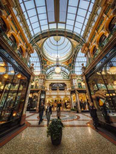 County Arcade in Leeds presents a sumptuous Victorian arcade of exceptional architectural refinement, its soaring barrel-vaulted glass roof supported by ornamental cast-iron ribs flooding the interior with soft, diffused natural light that illuminates the richly decorated terracotta and cream-coloured faience tilework adorning the vaulted ceiling. The corridor stretches away in symmetrical perspective, flanked on both sides by elegant shop frontages with mahogany-framed glazing and gilt detailing, their display windows glowing warmly beneath brass light fittings that cast pools of amber illumination across the tessellated terracotta and cream geometric floor patterning below. Ornamental green and gilt decorative panels and scrollwork embellish the walls between shopfronts, creating a rhythm of pattern and refinement typical of late-Victorian arcade design, whilst cream-coloured classical pilasters with decorative capitals provide structural and visual emphasis to the elevation. The arcade is animated with shoppers moving between the luxury retailers—visible signage includes Reiss amongst other boutique establishments—their silhouettes contrasting against the honey-toned architectural backdrop, whilst a potted ivy plant marks the central axis of the perspective, anchoring the composition with a touch of living green against the sumptuous architectural theatre surrounding it.