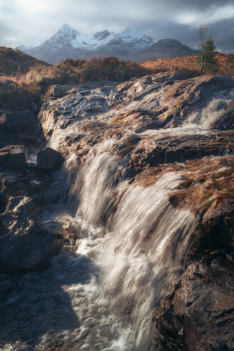 A beautiful scene from Sligachan on the Isle of Skye, Scotland, featuring a lively cascade of water flowing over dark, weathered rocks in the foreground. The light catches the water, giving it a silvery sheen as it tumbles down into a foamy pool. In the middle ground, rugged, orange-brown moorland rises gently, dotted with bushes and a solitary tree clinging to the rocky terrain. Towering in the background are dramatic, snow-capped peaks of the Black Cuillin mountains, partially shrouded in moody clouds, creating a striking contrast against the brighter foreground.