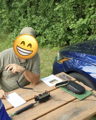 A person wearing a hat, face covered with a smiling emoji, holding the microphone to a small amateur radio on a picnic table in a park.